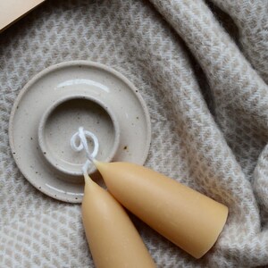 May include: Two tapered beeswax candles with white wicks on a white ceramic candle holder. The candles are on a beige and white patterned blanket.