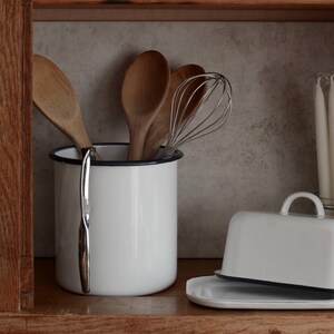May include: White enamel kitchenware with navy blue trim, including bowls, plates, and a mug. Wooden spoons, a whisk, and a butter dish are also visible. The items are arranged on a wooden shelf, creating a rustic, farmhouse aesthetic.