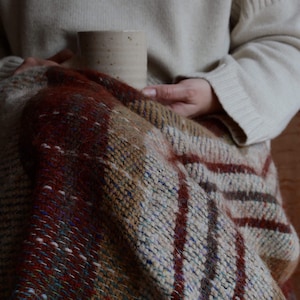 May include: A multi-coloured woven blanket, featuring red, brown, beige, and blue hues, is draped over someone. The blanket has a textured weave with vertical stripes and a fringed edge. A beige ceramic mug is held in the person's hands.