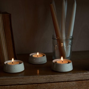 May include: Three lit tea light candles in speckled ceramic holders on a wooden surface. Tall, unlit taper candles stand in a glass jar in the background. A vintage book is partially visible on the left.