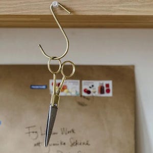 Peut inclure: Une paire de ciseaux dorés suspendus à un crochet doré. Les ciseaux sont suspendus au-dessus d'une enveloppe en papier brun avec du texte manuscrit.