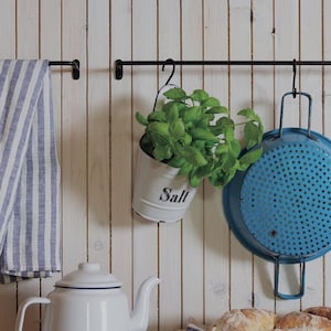 Może przedstawiać: Biały emaliowany czajnik z czarną obwódką, biały emaliowany pojemnik z napisem "Sól" na nim, wiszący na czarnym metalowym pręcie, niebieski durszlak wiszący na pręcie i drewniana miska wypełniona bułeczkami na drewnianym blacie.