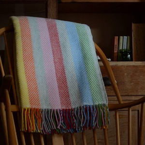 May include: A multi-coloured striped blanket draped over a wooden chair. The blanket has vertical stripes in yellow, orange, pink, red, blue, and green. The blanket has a fringe of multicoloured yarn at the bottom. The chair is made of light-coloured wood.