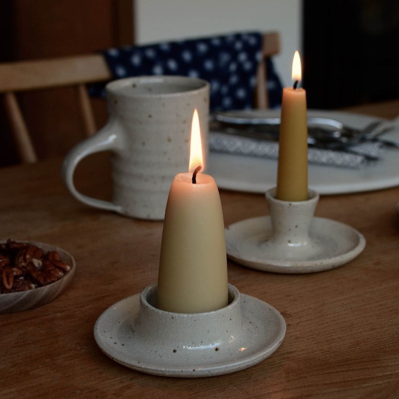 May include: Two white ceramic candle holders with a speckled finish. Each holder has a lit beige candle. The candles are on a wooden table.
