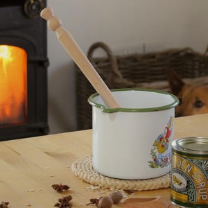 Peut inclure: Une casserole en émail blanc avec un bord vert et une cuillère en bois. La casserole a une illustration colorée et repose sur un dessous de plat tressé. Une boîte de Golden Syrup est à côté de la casserole.