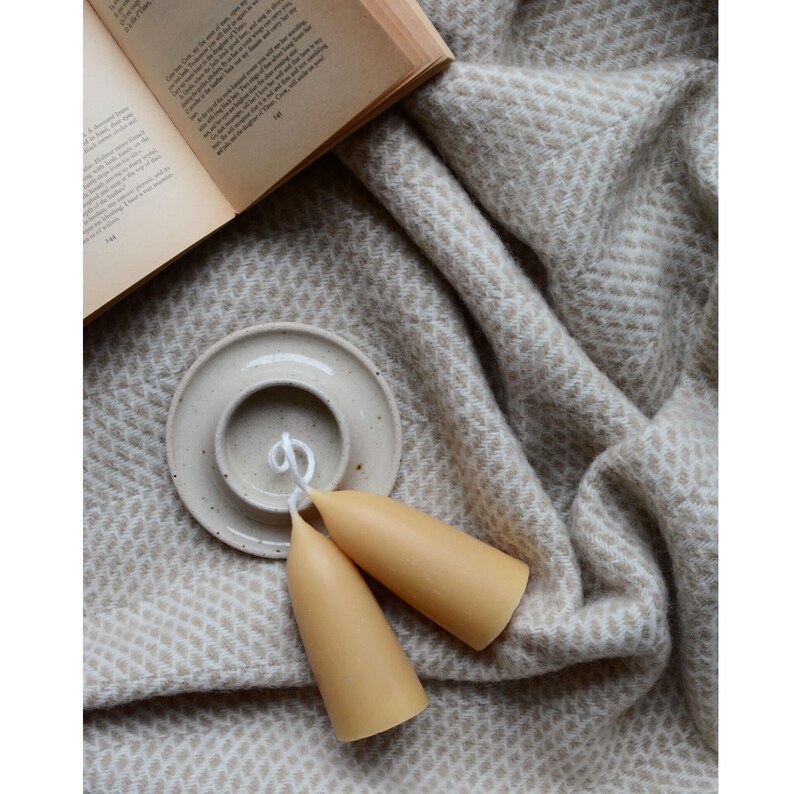 May include: Two tapered beeswax candles with white wicks on a white ceramic candle holder. The candles are on a beige and white patterned blanket.
