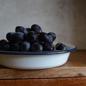 May include: A white enamel bowl with a dark blue rim filled with a cluster of dark purple grapes. The bowl is on a wooden surface, with a neutral background.