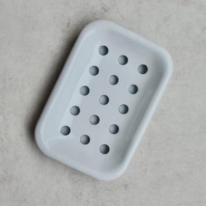 May include: A rectangular soap dish with a glossy light blue finish and rounded corners. The base of the dish features a grid of twelve circular drainage holes arranged in three rows of four, allowing for water to pass through the bottom surface.