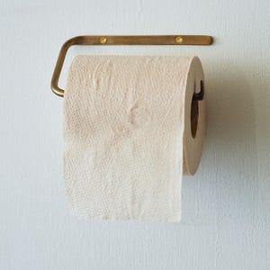 May include: A roll of beige toilet paper hangs on a brass-colored metal holder. The holder is mounted on a white wall. The toilet paper is textured and partially unrolled.