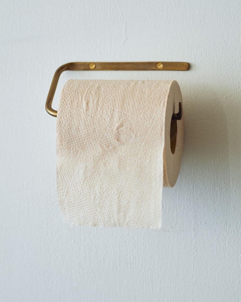 Pode incluir: Um rolo de papel higi&ecirc;nico bege em um suporte de metal cor de lat&atilde;o, montado em uma parede branca. O papel higi&ecirc;nico &eacute; texturizado e parcialmente desenrolado. O suporte tem um design simples.