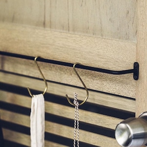 May include: A black metal bar with two brass hooks attached to a wooden wall. A white towel and a wooden utensil are hanging from the hooks.