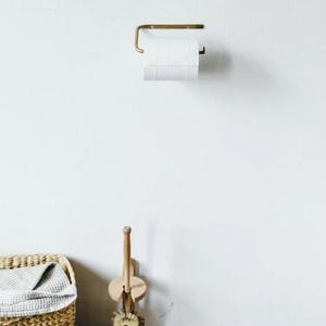 Pode incluir: Um porta-papel higi&ecirc;nico de cor lat&atilde;o com um rolo de papel branco est&aacute; montado em uma parede branca. Uma escova de vaso sanit&aacute;rio de madeira e uma cesta de vime com uma toalha cinza est&atilde;o no ch&atilde;o. Os itens est&atilde;o em um banheiro.