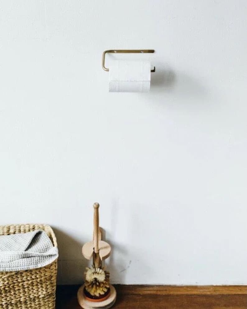 Pode incluir: Um porta-papel higi&ecirc;nico de cor lat&atilde;o com um rolo de papel branco est&aacute; montado em uma parede branca. Uma escova de vaso sanit&aacute;rio de madeira e uma cesta de vime com uma toalha cinza est&atilde;o no ch&atilde;o. Os itens est&atilde;o em um banheiro.
