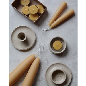 May include: A set of two natural beeswax taper candles and a set of six small round beeswax tea lights. The candles are on a white background with two ceramic candle holders.