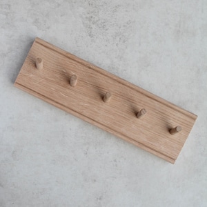 May include: A light-coloured wooden coat rack with five pegs. The rectangular rack has a decorative border and is made of natural wood. The pegs are evenly spaced along the length of the rack. The rack is shown on a light grey surface.
