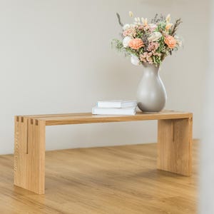 Könnte beinhalten: Eine helle Holzbank mit rechteckiger Oberseite und Beinen. Eine graue Vase mit einem Blumenstrauß und zwei weißen Büchern stehen auf der Bank. Die Blumen sind pfirsichfarben, weiß und grün. Die Bank ist etwa 100 cm lang.