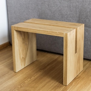 Könnte beinhalten: Ein heller Holzhocker mit schlichtem Design. Der Hocker hat eine rechteckige Oberseite und vier Beine. Der Hocker ist aus massivem Holz gefertigt und hat eine natürliche Oberfläche.
