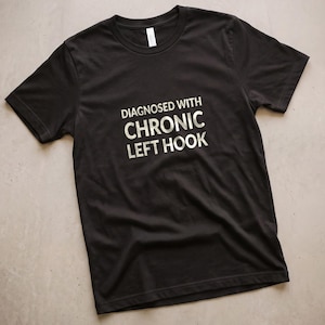 May include: A black short-sleeved t-shirt featuring the white text "DIAGNOSED WITH CHRONIC LEFT HOOK" printed in a bold, sans-serif font across the center of the chest. The garment has a crew neckline and a standard fit with visible fabric folds.