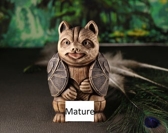 May include: A wooden figurine of a raccoon wearing a shell, with the word "Mature" written on a white tag in front of it. The figurine is sitting on a brown surface with a peacock feather behind it.