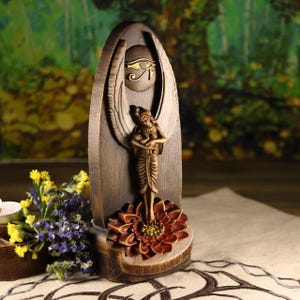 K&ouml;nnte beinhalten: Holzskulptur einer gefl&uuml;gelten Frau, die auf einer Lotusblume steht. Die Skulptur hat eine dunkelbraune Oberfl&auml;che mit goldenen Akzenten, einschlie&szlig;lich des Horusauges. Der Hintergrund ist verschwommen, mit Andeutungen von Gr&uuml;n und Braun. Handgeschnitzte Skulptur.