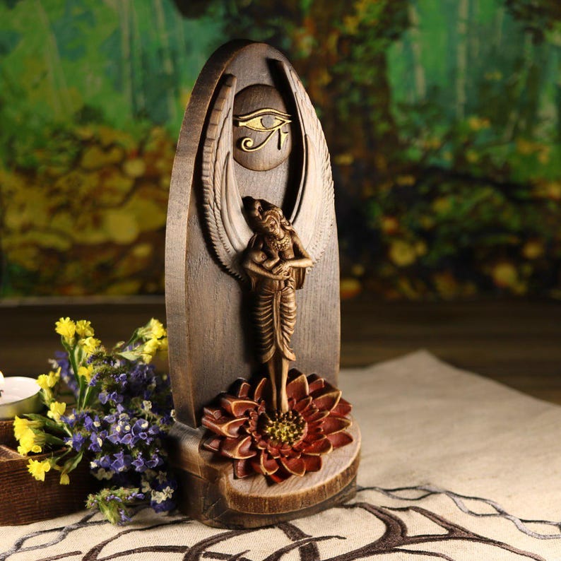 K&ouml;nnte beinhalten: Holzskulptur einer gefl&uuml;gelten Frau, die auf einer Lotusblume steht. Die Skulptur hat eine dunkelbraune Oberfl&auml;che mit goldenen Akzenten, einschlie&szlig;lich des Horusauges. Der Hintergrund ist verschwommen, mit Andeutungen von Gr&uuml;n und Braun. Handgeschnitzte Skulptur.