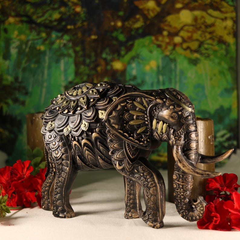 May include: A decorative elephant figurine with intricate details and gold accents. The elephant is standing on a white surface with red flowers in the background.