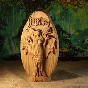 May include: A carved wooden sculpture of a female figure with dragon wings and two dragon heads. The figure is wearing a long dress and holding objects. The word "Bune" is carved at the base, with text at the top of the sculpture.