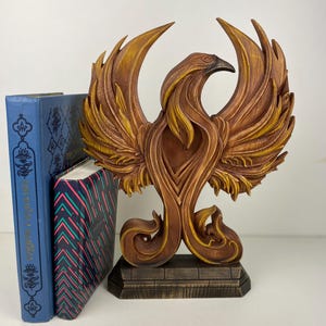 Op de afbeelding: Gedetailleerde houten fenikssculptuur met uitgespreide vleugels, in bruin- en goudtinten. De mythische vogel staat op een rechthoekige basis, geflankeerd door twee boeken. Een decoratief item voor in huis.