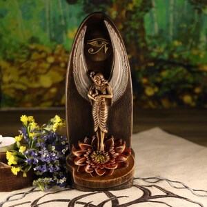 K&ouml;nnte beinhalten: Eine braune und goldene Statue einer Engelsfigur mit Fl&uuml;geln, die auf einer Lotusbl&uuml;tenbasis steht. Die Statue hat ein Augensymbol &uuml;ber dem Engel. Der Hintergrund ist verschwommen mit Gr&uuml;n- und Braunt&ouml;nen.
