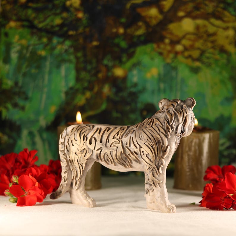 May include: A detailed ceramic tiger figurine with intricate carvings, standing on a white surface with red flowers and candles in the background.