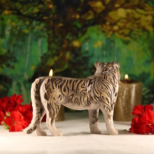 May include: A detailed, intricately carved tiger figurine made of a light brown material. The figurine is standing on a white surface with red flowers and candles in the background.