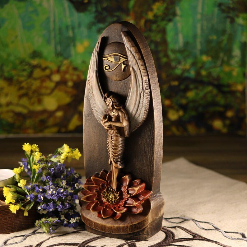 K&ouml;nnte beinhalten: Holzskulptur im &auml;gyptischen Stil. Sie zeigt eine gefl&uuml;gelte Figur, die auf einer Blume steht, mit einem Augensymbol dar&uuml;ber. Die Skulptur ist in Braun-, Gold- und Rott&ouml;nen gehalten, wahrscheinlich ein Dekorationsgegenstand.