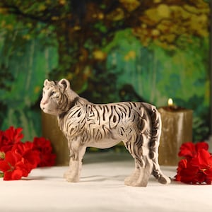 May include: A white ceramic tiger figurine with intricate details, standing on a white surface with red flowers in the foreground.