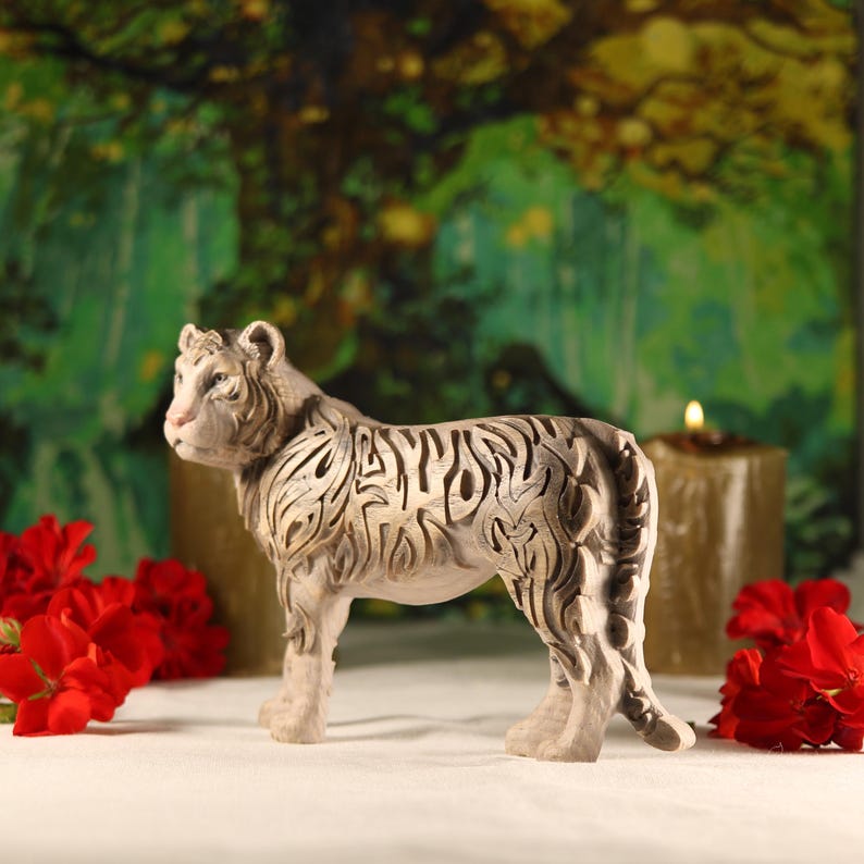 May include: A white ceramic tiger figurine with intricate details, standing on a white surface with red flowers in the foreground.