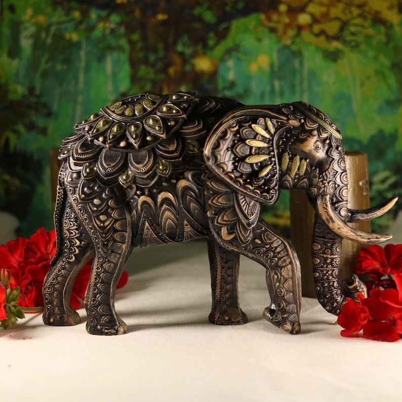 May include: A black and gold elephant figurine with intricate carvings and a decorative design on its back. The elephant is standing on a white surface with red flowers in the background.