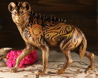 Puede incluir: Una figurita de hiena de madera con intrincados tallados de sol y motivos florales en tonos marrones y dorados. La figurita está de pie sobre una superficie de tela beige.