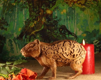 May include: A detailed, brown capybara sculpture with intricate carved patterns on its back. A lit red candle and orange roses are next to the sculpture. The background features a painted forest scene.