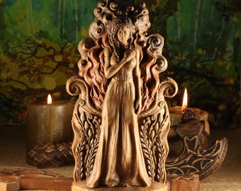May include: A wooden statue of a woman in a long dress, with intricate carvings and a flame-like design behind her. The statue is on a base with runes. Two lit candles and other wooden carvings are in the background.