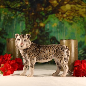May include: A white ceramic tiger figurine with intricate black and brown markings. The tiger is standing on a white surface with red flowers in the background.