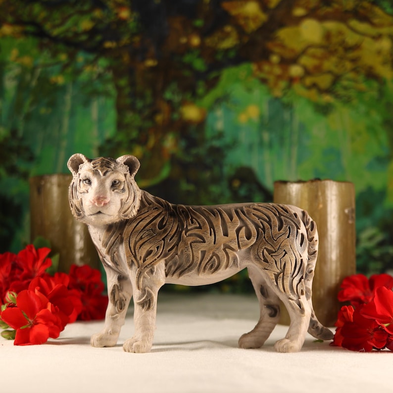 May include: A white ceramic tiger figurine with intricate black and brown markings. The tiger is standing on a white surface with red flowers in the background.