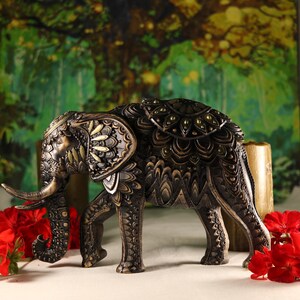 May include: A black and gold elephant figurine with intricate carvings and decorative details. The elephant is standing on a white surface with red flowers in the foreground.