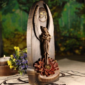 K&ouml;nnte beinhalten: Holzskulptur einer &auml;gyptischen Gottheit mit Fl&uuml;geln, die auf einer Lotusblume steht. Die Skulptur ist dunkelbraun mit goldenen Akzenten, einschlie&szlig;lich des Horusauges, gehalten. Der Hintergrund ist verschwommen, was eine nat&uuml;rliche Umgebung suggeriert.