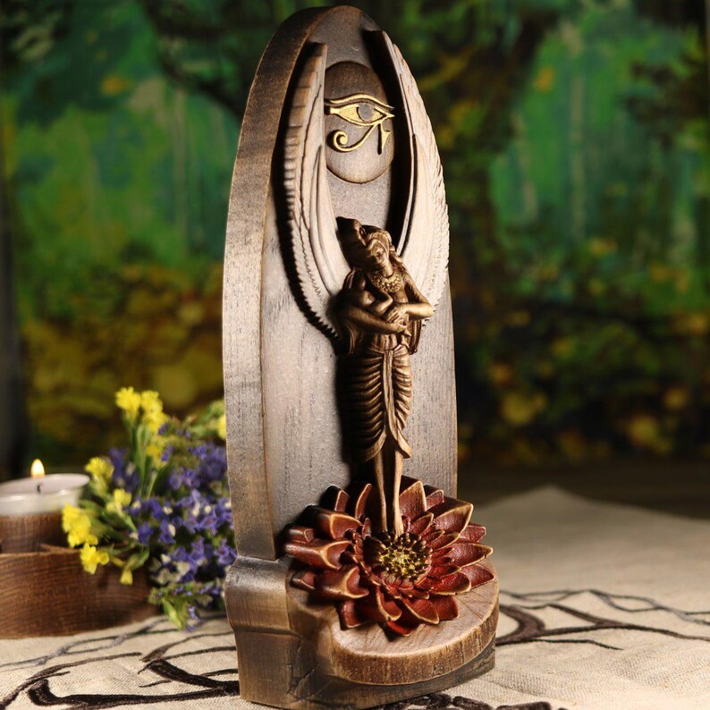 K&ouml;nnte beinhalten: Holzskulptur einer &auml;gyptischen Gottheit mit Fl&uuml;geln, die auf einer Lotusblume steht. Die Skulptur ist dunkelbraun mit goldenen Akzenten, einschlie&szlig;lich des Horusauges, gehalten. Der Hintergrund ist verschwommen, was eine nat&uuml;rliche Umgebung suggeriert.