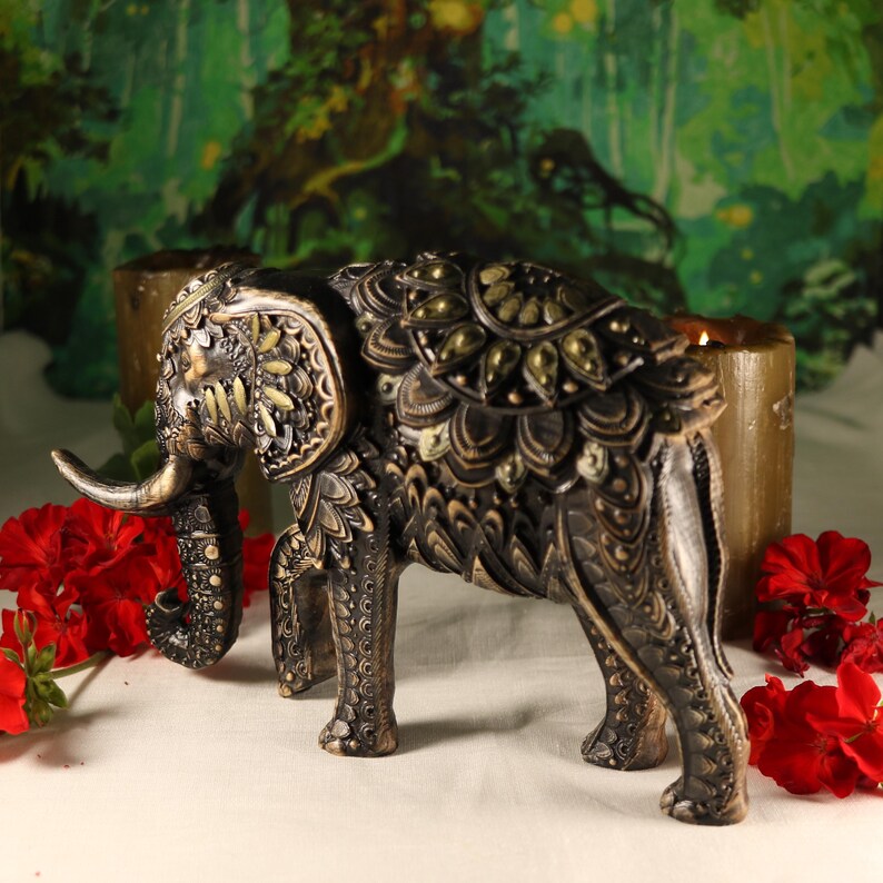 May include: A detailed, intricately carved elephant figurine with gold accents. The elephant is standing on a white surface with red flowers in the background.