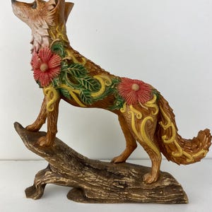 Könnte beinhalten: Eine braune Fuchsskulptur mit floralen und Ranken-Details. Der Fuchs steht auf einem Baumstamm, mit roten Blumen und grünen Blättern, die seinen Körper schmücken. Der Fuchs blickt nach oben, mit detaillierter Fellstruktur.