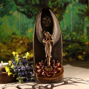 K&ouml;nnte beinhalten: Eine bronzefarbene Statue mit &auml;gyptischem Thema. Die Statue zeigt eine gefl&uuml;gelte Figur, die auf einer Lotusblume steht, mit einem Augensymbol dar&uuml;ber. Der Hintergrund ist eine dunkelbraune Holztafel. Die Statue ist ein Dekorationsartikel.