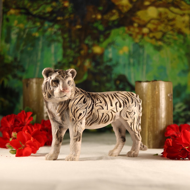 May include: A white ceramic tiger figurine with intricate carvings on its fur. The figurine is standing on a white surface with red flowers and brown candles in the background.