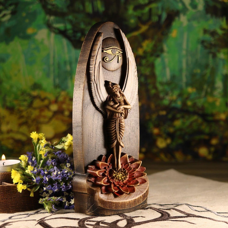 K&ouml;nnte beinhalten: Eine Holzskulptur mit einer Engelsfigur, die auf einer Lotusblume steht. Die Skulptur hat eine hellbraune Oberfl&auml;che mit einem goldfarbenen Horusauge-Symbol. Der Hintergrund ist ein verschwommenes Bild eines Waldes.