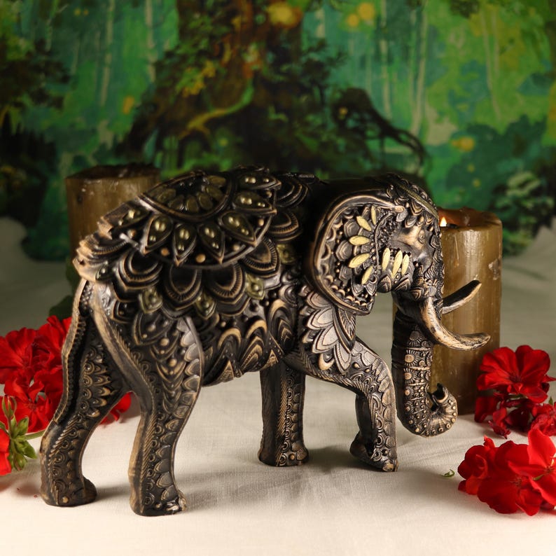 May include: A black and gold decorative elephant figurine with intricate carvings and details. The elephant is standing on a white surface with red flowers.