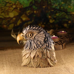 Puede incluir: Una detallada escultura de madera de la cabeza de un águila, con intrincadas tallas y una paleta de colores naturales. La escultura muestra el pico, los ojos y las plumas del ave, con un enfoque en la textura y la artesanía. La cabeza de águila es un objeto decorativo.